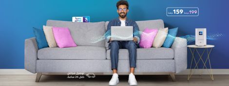 رجل يجلس على أريكة رمادية مع كمبيوتر محمول ووسائد ملونة على خلفية زرقاء